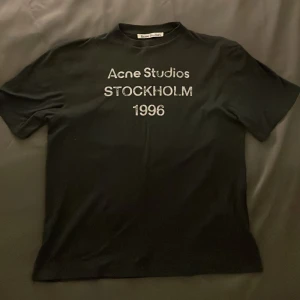 Svart Acne Studios t-shirt - Svart t-shirt från Acne Studios med tryck 'Acne Studios STOCKHOLM 1996' på bröstet. Klassisk passform och rund hals. Tillverkad i 85% bomull och 15% hampa för en mjuk och skön känsla. Perfekt för dig som gillar stilrena och trendiga plagg. Oversized fit sitter riktigt snyggt på kroppen. 