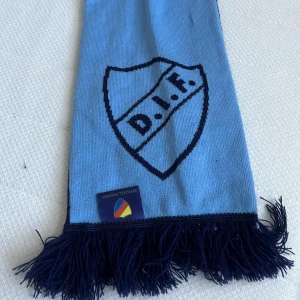 Djurgården halsduk blå och marinblå, aldrig använd - Säljer en lång supporterhalsduk från Djurgården med DIF-logga och texten 'Djurgården' samt 'Stockholmslaget' i blått och marinblått. Halsduken har fransar i ändarna och är stickad i mjukt material, perfekt för matchdagar och att visa vilket lag du håller på.