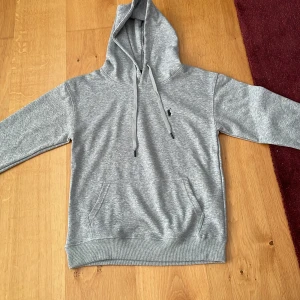 Grå hoodie från Polo Ralph Lauren - Snygg grå hoodie från Polo Ralph Lauren med klassisk broderad logga på bröstet. Tröjan har huva med snörning och en stor magficka framtill. Perfekt för en chill och stilren look. Tröjan är bara testad med lappen kvar på.