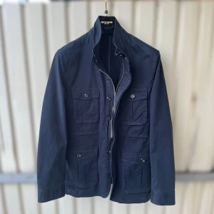 Marinblå FieldJacket från Gutteridge - Snygg marinblå jacka från Gutteridge med fyra stora fickor framtill, både knappar och dragkedja. Jackan har hög krage och stilren design, perfekt för dig som gillar klassisk look. Materialet är bomull och den har extra knappar med på etiketten.