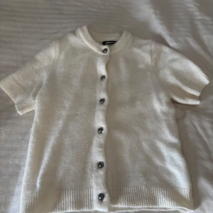 Vit stickad kofta från Gina Tricot - Kortärmad vit stickad kofta från Gina Tricot med rund hals och snygga silvriga knappar framtill. Mjuk ribbad kant vid ärmslut och nederkant, perfekt för lager-på-lager. Enkel och clean design som funkar till många stilar.