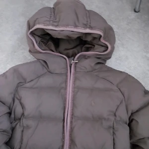 Brun dunjacka från Ralph Lauren - Säljer en brun dunjacka från Ralph Lauren med huva och dragkedja framtill. Jackan har quiltad design och diskret logga på bröstet. Materialet är polyester och nylon, vilket gör den både lätt och varm. Perfekt för kalla dagar och har en snygg loose passform.