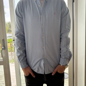 Rutig blåvit skjorta Polo Ralph Lauren - Säljer en slim fit skjorta från Polo Ralph Lauren i blåvitt rutigt mönster. Skjortan har klassisk krage med knappar, lång ärm och broderad rosa logga på bröstet. Tillverkad i mjuk bomull och passar perfekt till jeans eller chinos.