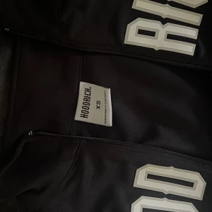 Svart hoodie från Hoodrich med vit text - Svart hoodie från Hoodrich med vit text framtill och vit rand längs ärmarna. Tröjan har dragkedja hela vägen, hög krage och en sportig vibe. Perfekt för dig som gillar streetwear och vill sticka ut med en snygg logga.