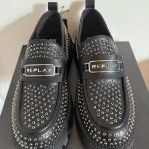 Svarta loafers med nitar från Replay - Chunky svarta loafers från Replay med grov platt sula och massor av silvriga nitar över hela skon. Skorna har Replay-logga framtill och coola detaljer i metall. Tillverkade i skinn och har en edgy vibe som sticker ut.