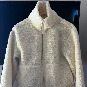 Vit teddyjacka med dragkedja, S - Mysig vit teddyjacka i storlek S med hög krage och hel dragkedja framtill. Jackan har en fluffig och mjuk känsla, perfekt för kyliga dagar. Enkel och stilren design som passar till det mesta. Helt slutsåld på H&M