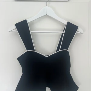 Svart topp från Club Monaco - Supersnygg svart topp från Club Monaco med breda axelband och vita kontrastsömmar. Toppen har en figurnära passform. Materialet är en mix av viskos och polyester som ger en lyxig känsla.