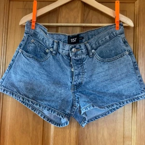 Blå jeansshorts med broderade fickor - Snygga blå jeansshorts från 157 med hög midja och klassisk femficksdesign. Baksidan har två fickor med coolt svart broderat mönster. Shortsen har knapp och dragkedja framtill och är gjorda i slitstarkt denim. Perfekta för dig som gillar en chill och trendig stil.