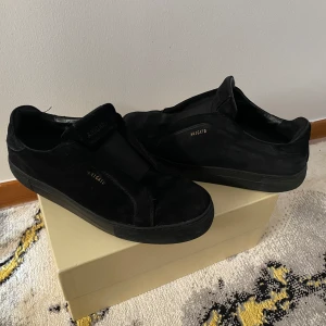 Svarta sneakers Axel Arigato mocka - Stilrena svarta sneakers från Axel Arigato i mjuk mocka med diskret logga i guld på sidan. Skorna har en minimalistisk design utan snörning, med elastiskt band över vristen och platt sula. Perfekta för dig som gillar en clean och modern look.