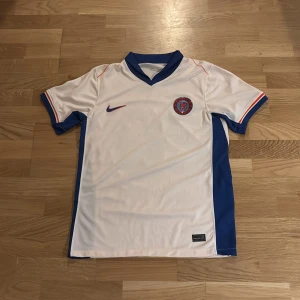 Chelsea Palmer 20 bortatröja Nike M - Chelsea FC bortatröja med Palmer 20-tryck. Vit med blåa och orange detaljer, blå krage och ärmslut. Tillverkad i Nike Dri-FIT-material som andas. Klassisk passform och officiellt klubbmärke på bröstet. Perfekt för dig som vill representera Chelsea på match eller i vardagen.  100% polyester