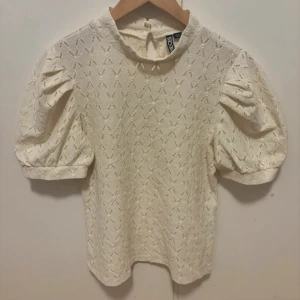 Vit blus med puffärm från Pieces - Supersöt vit blus från Pieces med puffiga korta ärmar och broderat hålmönster. Blusen har hög hals med knapp i nacken och en lätt strukturerad känsla. Perfekt för dig som gillar trendiga detaljer. Frakt ingår ej💕
