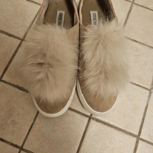 Beige sneakers med fluff från Steve Madden - Snygga slip-on sneakers från Steve Madden i beige mocka med vit sula och stora fluffiga pälsdetaljer på ovansidan. Skorna har en rund tå och platt sula, perfekt för dig som vill sticka ut med en unik och trendig look.