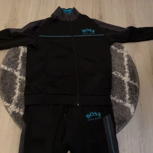 Svart ziptröja från Hugo Boss och byxor  - Svart ziptröja från Hugo Boss med blå logga på bröstet och tunna blå ränder över bröstet. Tröjan har hög krage, hel dragkedja och långa ärmar. Perfekt för en sportig och stilren look samt byxor till. Priset är inte hugget i sten.