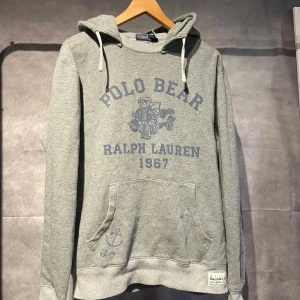 Ralph Lauren hoodie  - Grå hoodie från Polo Ralph Lauren med blå Polo Bear-tryck och text framtill. Tröjan har huva med snörning, känguruficka och ribbade muddar. Mjuk bomullskvalitet och cool detalj med patch nertill. Perfekt för chill dagar och streetstyle.