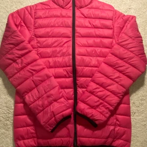 Rosa pufferjacka i storlek 42/44 - Säljer en snygg rosa pufferjacka med horisontella sömmar och hög krage. Jackan har hel dragkedja framtill och är lätt vadderad, perfekt för kyliga dagar. Passformen är normal och materialet känns som syntet. Enkel och stilren design utan synligt märke.