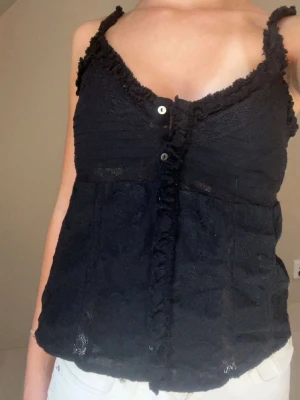 Svart spetslinne med knappar - Säljer ett svart linne med spetsdetaljer och dekorativa knappar framtill. Linnet har smala axelband och volangkant längs urringningen. Materialet är spets med blommönster och linnet är lätt croppad.