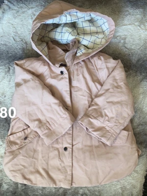 Ljusrosa vinterjacka med huva - Säljer en ljusrosa vinterjacka med huva och rutigt foder. Jackan har knappar framtill och två fickor med lock. Perfekt för kalla dagar och enkel att matcha till olika outfits.🌸 Fint använt skick. Storlek 80