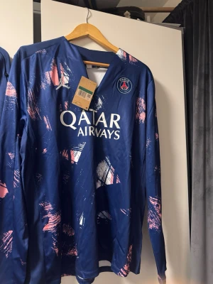 PSG träningsset blå/rosa Jordan - Säljer ett fett PSG träningsset med långärmad tröja och byxor i marinblått med rosa och vita detaljer. Tröjan har Paris Saint-Germain-logga, Qatar Airways-tryck och coolt mönster. Byxorna har elastisk midja och PSG-märke. Perfekt för fotbollsträning eller chill.