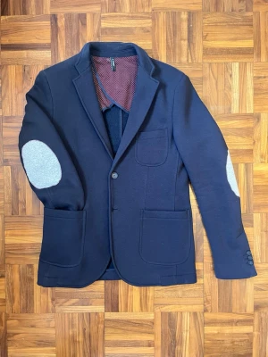 Marinblå kavaj från A Deep - Snygg marinblå kavaj från A Deep, tillverkad i Italien. Jackan har ljusgrå armbågslappar, tre framfickor och knappar vid ärmslut. Insidan är fodrad med vinröd prickigt tyg som ger en cool kontrast. Perfekt för dig som vill sticka ut med stil. Kavaj i sweatshirt material 