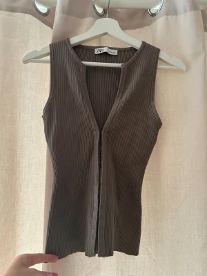 Brun ribbad linnetopp från Zara - Säljer en brun ribbad linnetopp från Zara i storlek S. Toppen är ärmlös med djup v-ringning och har en snygg knäppning framtill. Materialet är mjukt och stretchigt, perfekt för att styla med jeans eller kjol.