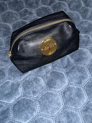 Svart Mulberry necessär i skinn - Säljer en svart necessär från Mulberry i äkta skinn med guldiga metalldetaljer och dragkedja. Väskan har Mulberrys klassiska logga framtill och en stilren, enkel form som passar till många olika looks. Perfekt för dig som gillar lyxiga accessoarer med tidlös känsla.