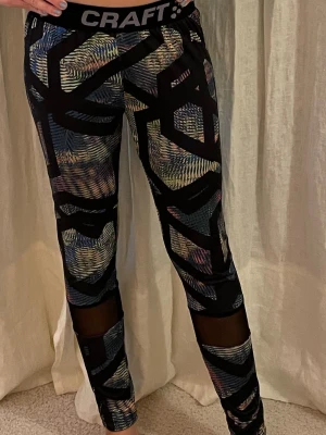 Craft leggings med mesh och mönster. - Säljer ett par snygga leggings från Craft med svart bas och färgglatt abstrakt mönster. De har bred resår i midjan och meshpaneler på benen för extra ventilation. 🩷