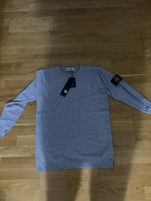 Grå stickad tröja från Stone Island - Säljer en stilren grå stickad tröja från Stone Island med klassisk rund hals och ikonisk patch på ärmen. Tröjan har ribbade muddar vid ärmslut och nederkant, perfekt för dig som gillar clean streetwear med premiumkänsla.