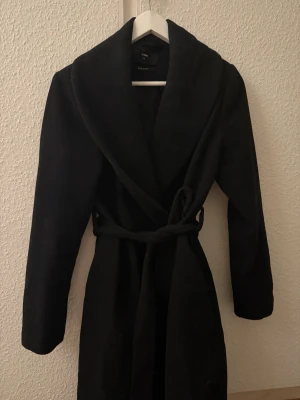 Svart kappa från BikBok - Säljer en svart kappa från BikBok i ullmix. Jackan har en snygg omlottmodell med brett slag och knyts i midjan med bälte. Långärmad och klassisk, perfekt för kyliga dagar. Enkel och stilren design som passar till allt. Köpt begagnad och kommer inte till användning