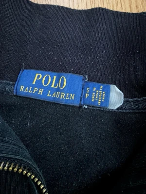 Svart half zip tröja Polo Ralph Lauren - Klassisk svart tröja från Polo Ralph Lauren med röd broderad logga på bröstet. Modellen har half zip-dragkedja och hög krage. Tillverkad i mjuk bomull och har långa ärmar. Perfekt för en chill och stilren look.