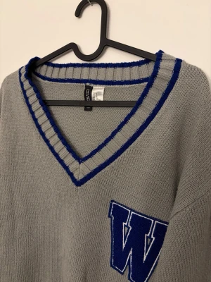 Grå stickad tröja med blå W - Collegeinspirerad grå stickad tröja från Divided med blåa ränder vid v-ringad hals, ärmslut och nederkant. Använd få gånger men kommer inte till användning 