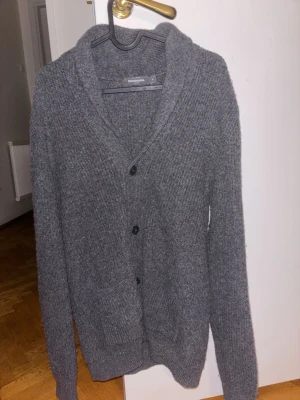 Grå stickad cardigan från Dressmann - Mysig grå stickad cardigan från Dressmann i storlek M. Koftan har 4 knappar framtill, två fickor och en bred krage. Perfekt för lager på lager och chill höst- eller vinterstil.