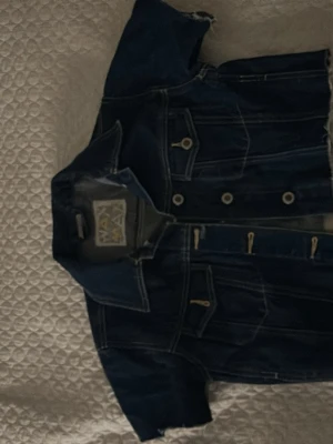 Mörkblå jeansjacka med stjärndetalj - Säljer en mörkblå jeansjacka från Wax Max med korta ärmar och råa kanter. Jackan har knappar framtill och en snygg stjärna av nitar på ryggen. Najs till sommarn! Är uttöjd så funkar på folk som har xs i storlek