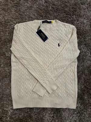 Vit tröja Polo Ralph Lauren - Kabelstickad vit tröja från Polo Ralph Lauren med klassisk rund hals och broderad logga på bröstet. Tillverkad i mjuk bomull och har långa ärmar samt ribbade muddar. Perfekt för en clean och stilren look.