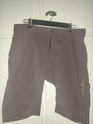 Bruna shorts från Houdini, storlek L - Säljer ett par bruna Houdini shorts i storlek L. De är tillverkade i ett lätt syntetmaterial och har en orange dragkedja på benet för extra förvaring. Perfekta för utomhusaktiviteter och sommarens äventyr. Vandringsshorts. 