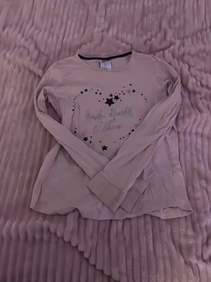 Ljusrosa långärmad tröja med stjärnor - Söt ljusrosa långärmad tröja med tryck av svarta och silvriga stjärnor i ett hjärtmönster på bröstet. Texten i mitten ger en extra gullig touch. Perfekt för dig som gillar mysiga och stilrena plagg med lite extra detaljer.