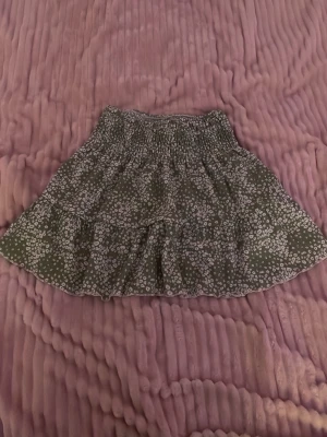 Grön blommig kortkjol med volang - Supersöt grön kortkjol med små vita blommor och volangdetaljer. Kjolen har en smockad midja som gör den bekväm och lätt att bära. Perfekt för dig som gillar en feminin och avslappnad stil.