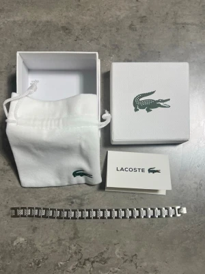 Snygg silverfärgad Lacoste armband - Stilrent armband från Lacoste i silverfärgad metall med klassiska krokodil-loggor på varje länk. Kommer i original ask och tygpåse med Lacoste-logga. Perfekt accessoar för dig som vill ha en clean och modern look.