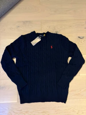 Mörkblå kabelstickad tröja Polo Ralph Lauren - Kabelstickad mörkblå tröja från Polo Ralph Lauren i storlek S. Klassisk modell med rund hals och röd broderad logga på bröstet. Tillverkad i mjuk bomull och har långa ärmar samt ribbade muddar. Perfekt för en clean och stilren look.