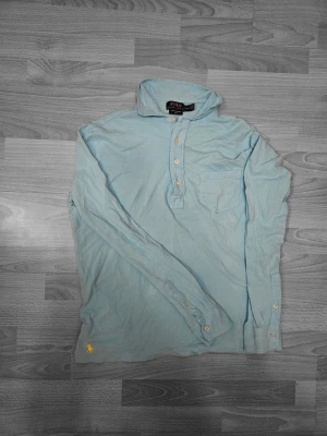 Ljusblå henley från Polo Ralph Lauren - Säljer en ljusblå henley från Polo Ralph Lauren i mesh-bomull. Skjortan har lång ärm, klassisk krage, knappar framtill och en bröstficka. Ikoniska gula Polo-loggan broderad nertill. Perfekt för dig som gillar stilrena och bekväma plagg.