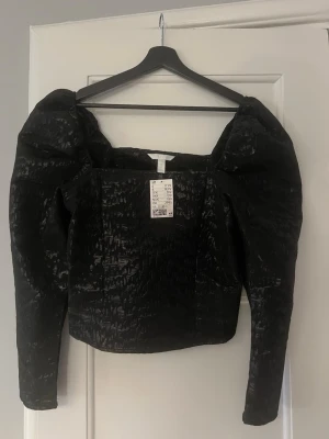 Svart puffärm blus från H&M - Säljer en svart blus från H&M med puffiga långa ärmar och fyrkantig urringning. Materialet har en glansig, instead look och ger en lyxig känsla. Perfekt för dig som vill sticka ut med coola detaljer och trendig siluett.
