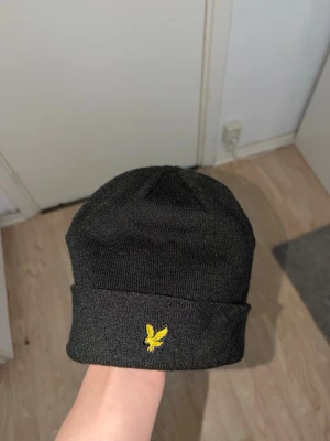 Lyle & scott mössa - svart mössa från lyle & scott med gul broderad logga framtill.