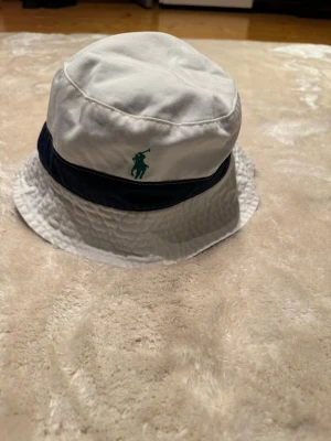 Vit och marinblå bucket hat Polo Ralph Lauren - Bucket hat från Polo Ralph Lauren i vit och marinblå bomull med grön broderad logga framtill. Insidan har ett blommigt mönster i blå och beige toner. Perfekt för att lyfta din stil med en cool detalj.