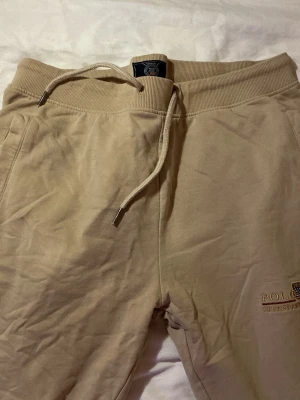 Beige mjukisbyxor från Vinson Polo Club - Beiga mjukisbyxor från Vinson Polo Club med broderad logga på benet. Byxorna har resår i midjan med snörning och mjukt, fleecefodrat material på insidan. Klassisk passform med fickor på sidorna och baktill. Perfekta för chill dagar.