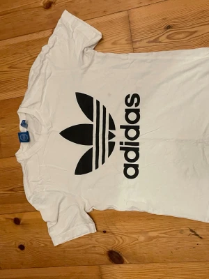 Vit Adidas t-shirt med stor logga - Säljer en vit t-shirt från Adidas med den klassiska svarta Trefoil-loggan tryckt stort på bröstet. T-shirten har rund hals och korta ärmar, tillverkad i mjuk bomull för skön känsla. Perfekt för dig som gillar sportig och clean stil. En liten fläck 
