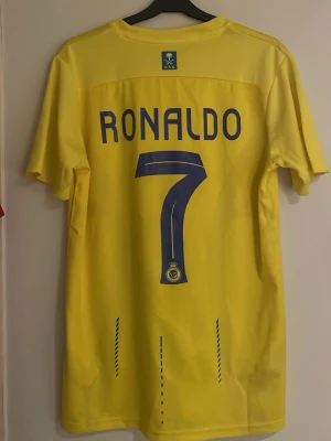 Gul Al Nassr Ronaldo matchtröja - Säljer en gul Al Nassr matchtröja med Ronaldo och nummer 7 på ryggen. Tröjan har blå detaljer, klubbmärke på bröstet och tryck framtill. Perfekt för dig som vill visa ditt stöd för laget och Ronaldo.