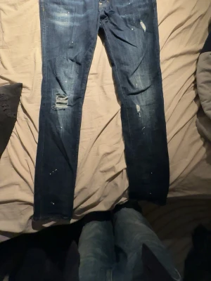 Mörkblå Dsquared2 jeans med slitningar - Säljer ett par mörkblå jeans från Dsquared2 med coola slitningar och färgstänk. Klassisk femficksmodell med gul kontrastsöm och stor röd Dsquared2-patch bak. Jeansen har rak passform och är tillverkade i robust denim. Perfekta för dig som gillar streetwear vibes.