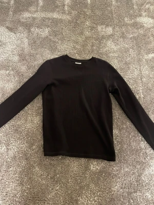 Svart ribbad långärmad topp - En enkel svart långärmad topp med ribbad struktur. Modellen har rund halsringning och är tillverkad i ett mjukt, stretchigt material som sitter skönt på kroppen. Perfekt att styla med jeans eller kjol för en clean look.