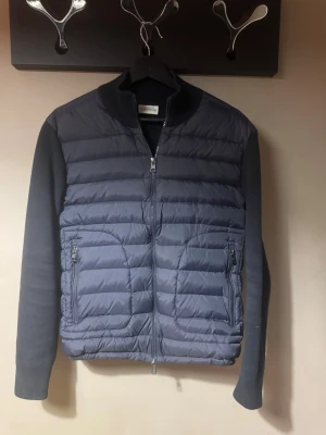 Mörkblå Moncler Cardigan jacka strl M - Säljer en mörkblå / navy snygg cardigan från Moncler med quiltad framsida och släta stickade ärmar och rygg. Jackan har två fickor med dragkedja och hög krage. Väldigt stilren, knappt använd och i väldigt bra skick. Jackan är i storlek M men skulle även säga att den passar S. Perfekt för dig som vill ha något stilrent och bekvämt.