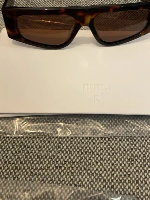 Brunmönstrad solglasögon från Filippa K - Snygga solglasögon från Filippa K i brunmelerat mönster. Glasögonen har en klassisk rak passform och är tillverkad i ett stilrent material som passar perfekt till lager-på-lager. Perfekt för dig som vill ha en trendig och tidlös look.