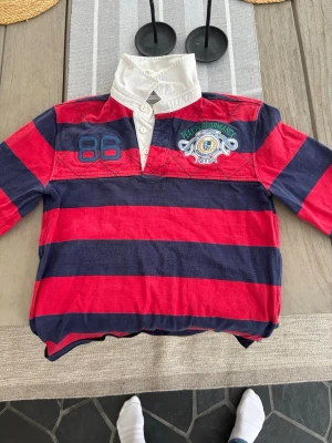 Randig pikétröja Peak Performance L - Säljer en klassisk randig pikétröja från Peak Performance med breda röda och marinblå ränder, vit krage och broderad logga på bröstet. Tröjan har lång ärm, knappar i halsen och coola detaljer som siffran 86 och brodyr. Perfekt för dig som gillar sportig stil! Bra skick och i storlek L